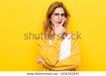young pretty blonde woman looking serious, thoughtful and distrustful, with one arm crossed and hand on chin, weighting options against yellow wall