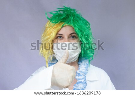 Woman in green and yellow wig, with mask and protective vestments against the coronavirus, makes a positive sign with her hands (thumbs up). White background.