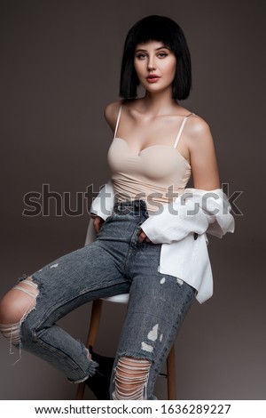 young beautiful brunette with short haircut. model wearing white jacket and jeans. lady in casual clothes posing on grey studio background. model on bar chair