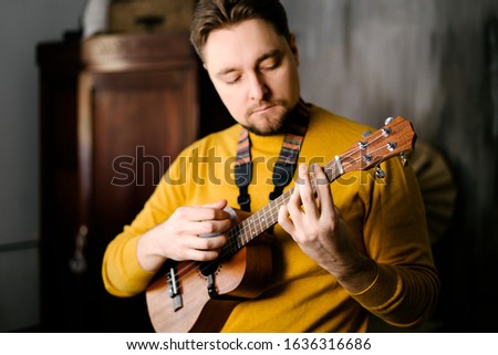 
A man with a beard and a yellow sweater plays the ukulele. Male musician with ukulele posing for the camera Royalty-Free Stock Photo #1636316686