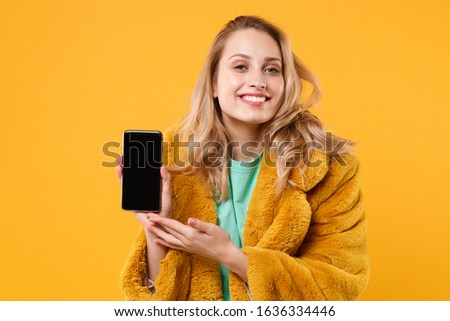 Smiling young blonde woman girl in yellow fur coat posing isolated on orange wall background studio portrait. People lifestyle concept. Mock up copy space. Hold mobile phone with blank empty screen