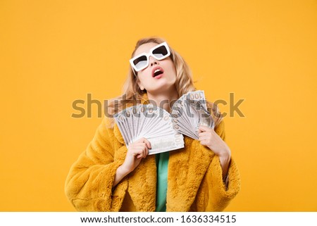 Stunning young blonde woman girl in yellow fur coat, dark sunglasses isolated on orange wall background. People lifestyle concept. Mock up copy space. Holding fan of cash money in dollar banknotes