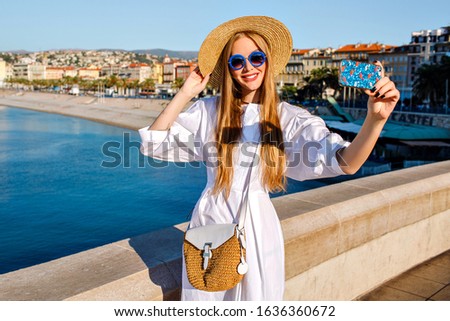 Elegant glamour magnificent woman wearing luxury white dress and straw accessories making selfie at beach front in French Riviera, traveling goals, bright sunny colors, blue ocean.