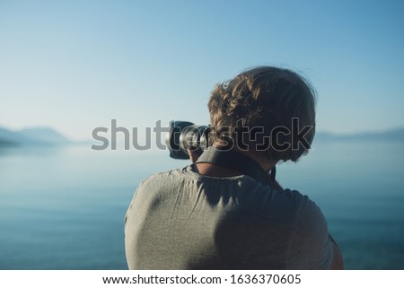 View from behind of a young male professional photographer taking a photo of a distant island as he stand by the ocean in the morning.
