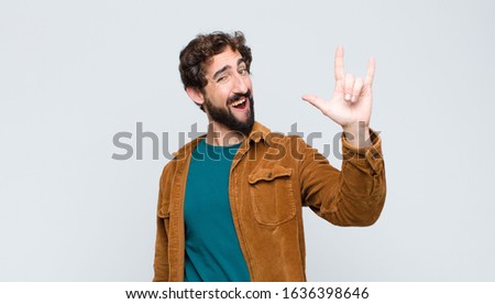 young handsome man feeling happy, fun, confident, positive and rebellious, making rock or heavy metal sign with hand against flat wall