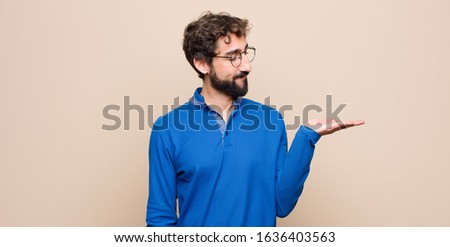 young handsome man feeling happy and smiling casually, looking to an object or concept held on the hand on the side against flat wall