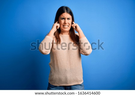 Young beautiful woman wearing casual sweater over blue background covering ears with fingers with annoyed expression for the noise of loud music. Deaf concept.