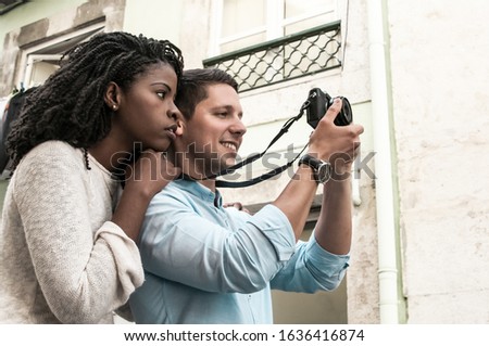 Positive interracial couple taking photos in city. Woman standing and leaning on man shoulder in street with old building in background. Romance and tourism concept. Side view.