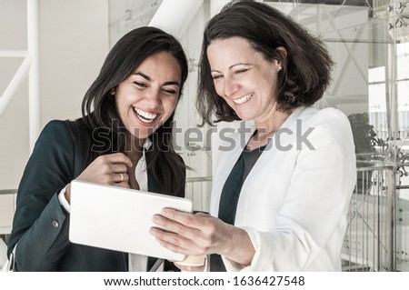 Two smiling business women using tablet in office hall. Colleagues or partners standing and videoconferencing or taking selfie photo. Technology and break concept. Front view.