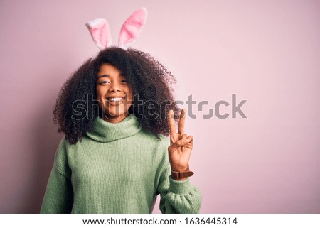 Young african american woman with afro hair wearing easter rabbit ears costume over pink background smiling with happy face winking at the camera doing victory sign. Number two.