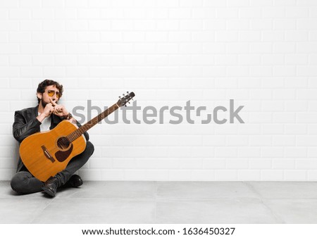 young musician man looking serious and displeased with both fingers crossed up front in rejection, asking for silence with a guitar, rock and roll concept