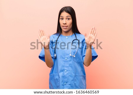 young latin nurse looking nervous, anxious and concerned, saying not my fault or I didn‚Äôt do it against pink wall