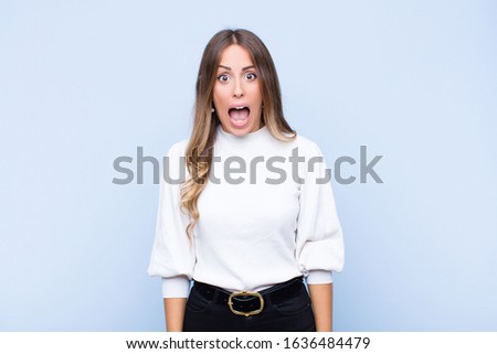 young pretty hispanic woman feeling terrified and shocked, with mouth wide open in surprise against blue wall