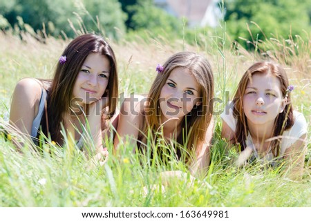 Three happy smiling & looking at camera teenage girl friends lying in high green grass on summer outdoors background