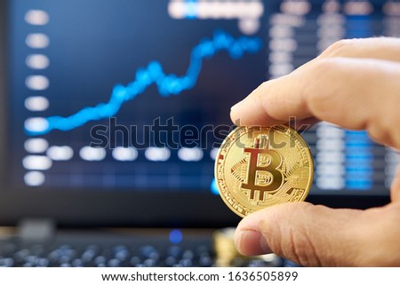 Male hand showing gold bitcoin. laptop keyboard with basic candlestick green red graph price in background. Worldwide cryptocurrency and digital payment concepts. Bitcoin price rises