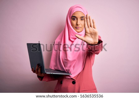 Young beautiful businesswoman wearing pink muslim hijab and business jacket using laptop with open hand doing stop sign with serious and confident expression, defense gesture