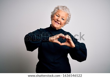 Senior beautiful woman wearing casual black sweater standing over isolated white background smiling in love doing heart symbol shape with hands. Romantic concept.