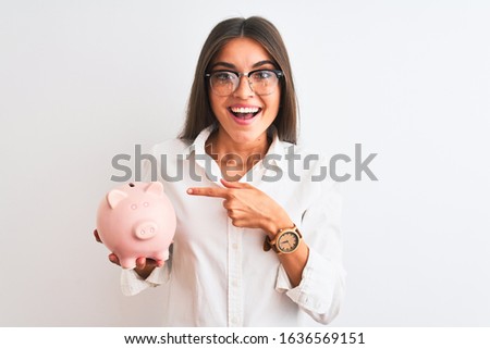 Beautiful businesswoman wearing glasses holding piggy bank over isolated white background very happy pointing with hand and finger