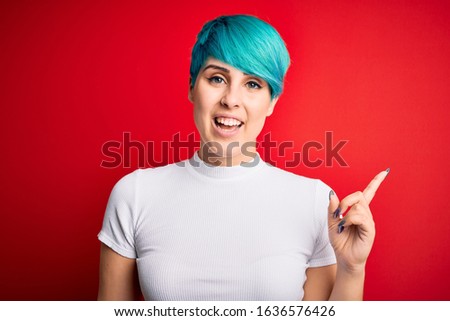 Young beautiful woman with blue fashion hair wearing casual t-shirt over red background with a big smile on face, pointing with hand finger to the side looking at the camera.