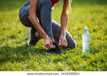 Cropped picture of caucasian woman in sportswear kneeling on meadow and tying shoelace on sneaker. Next to her is bottle of water. Sunny morning in nature.