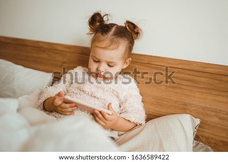 Happy little girl watching cartoons on phone lying In bed at home. Happy loving family. Modern wireless tech usage free time concept. Bedtime Fun.