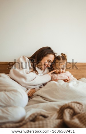  Cheerful mother and her daughter little daughter watching cartoons on phone lying In bed at home. Happy loving family. Modern wireless tech usage free time concept. Bedtime Fun.