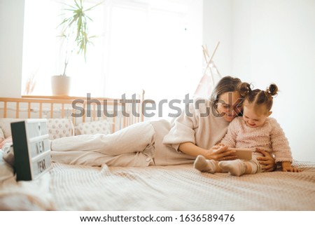  Cheerful mother and her daughter little daughter watching cartoons on phone lying In bed at home. Happy loving family. Modern wireless tech usage free time concept. Bedtime Fun.