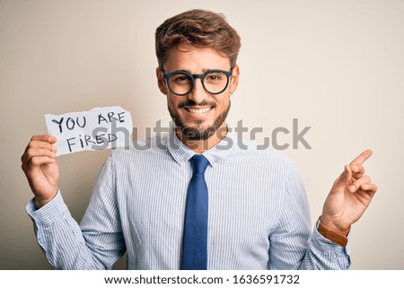 Young businessman wearing glasses holding paper with you are fired message very happy pointing with hand and finger to the side