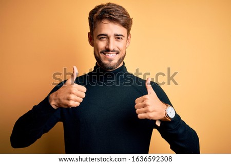 Young handsome man with beard wearing turtleneck sweater standing over yellow background success sign doing positive gesture with hand, thumbs up smiling and happy. Cheerful expression and winner 