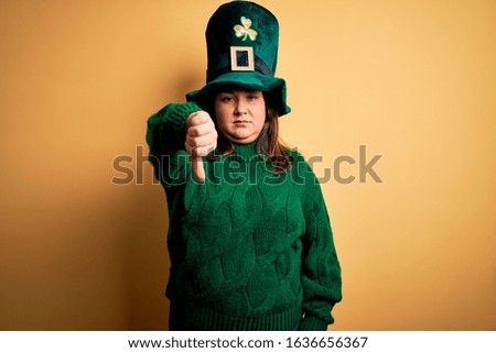 Young beautiful plus size woman wearing green hat with clover celebrating saint patricks day looking unhappy and angry showing rejection and negative with thumbs down gesture. Bad expression.