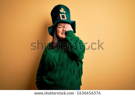 Young beautiful plus size woman wearing green hat with clover celebrating saint patricks day covering one eye with hand, confident smile on face and surprise emotion.