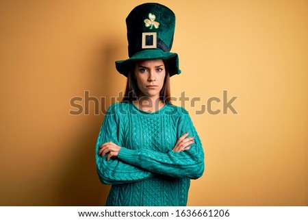 Young beautiful brunette woman wearing green hat with clover celebrating saint patricks day skeptic and nervous, disapproving expression on face with crossed arms. Negative person.