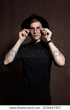 Elegant young man in round glasses, a black shirt with a hat on a brown background