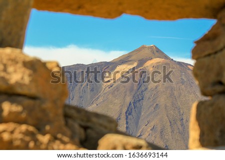 The picture shows the volcano from tenerife which is part of the canary islands from another mountain top throughout a windows made of rocks. It is a very sunny day in summer.