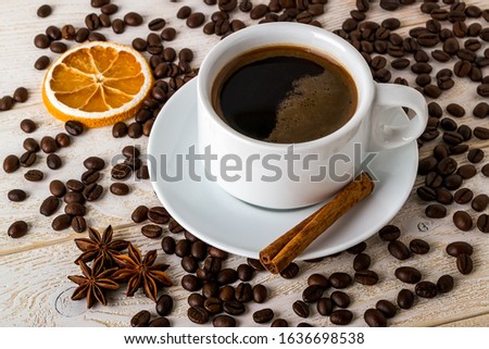 Hot coffee in white elegant cup with saucer on a background of coffe beans over a white wooden table. Coffee with cinnamon, anise and dry citrus. Front view.