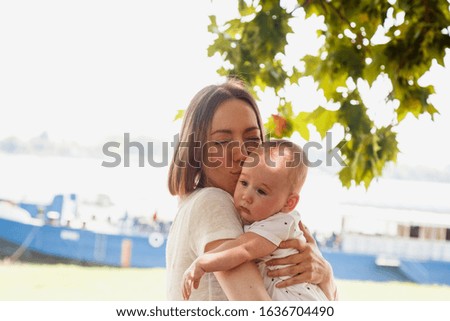 Happy slavic mother is hugs and kissing her newborn baby boy son outdoors near the river, bay area, under maple trees at the sunny day with boats at background.