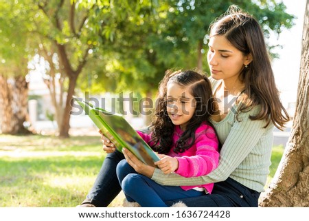 Young Mother Reading Picture Book To Daughter While Sitting Against Tree At Park