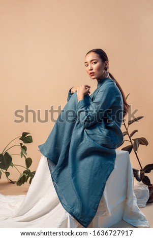 Young woman with ponytail wearing long dress sitting on white cube isolated on bage background with plants posing looking camera confident side view