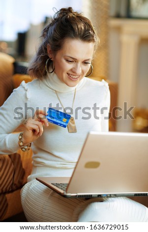 smiling modern woman in white sweater and skirt with credit card making online shopping on e-commerce website in the modern living room in sunny winter day.