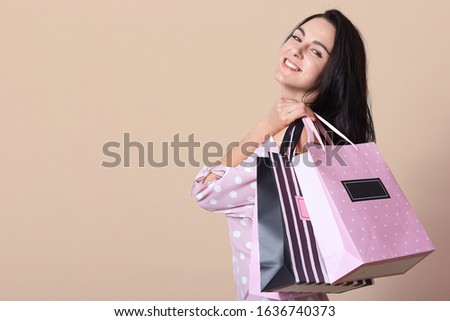 Picture of smiling sweet beautiful young female standing isolated over beige background in studio, holding light packages in one hand, looking directly at camera. Copyspace for advertisement.