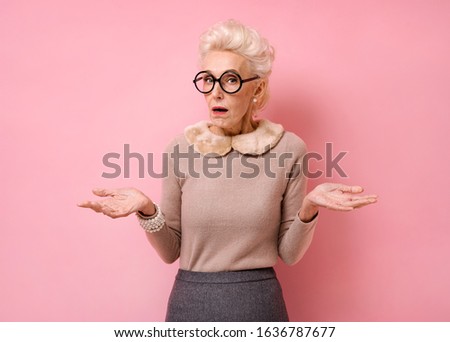Confused beautiful grandmother spreads hands sideways, feels doubt while makes choice. Photo of kind elderly woman in eyeglasses looking at camera on pink background.