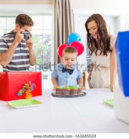 Father taking picture of birthday boy blowing out candles on cake at home