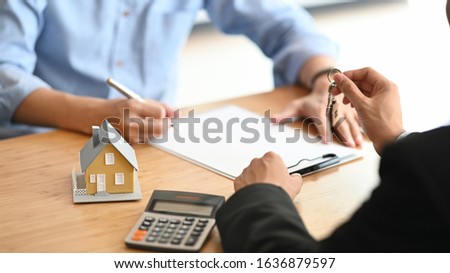 Cropped shot of House broker giving a property key to his customer while he has writing/signing on agreement at the modern wooden desk. Loan, Debt, Credit, Buying or Selling agreement concept.