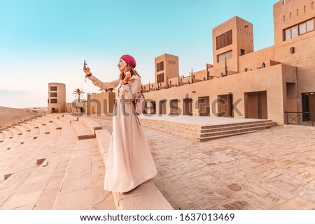 Happy woman traveler wearing dress and turban taking photos on her smartphone of an old Arab town or village in the middle of the desert. Concept of tourism and adventures