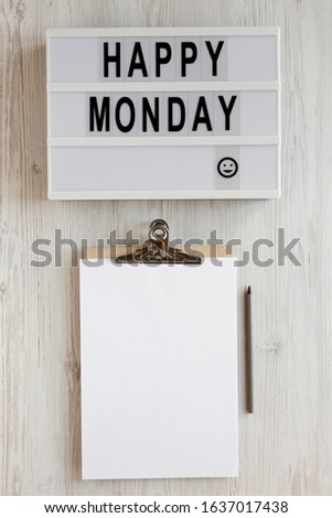 'Happy monday' words on a modern board, clipboard with blank sheet of paper on a white wooden surface, top view. Overhead, from above, flat lay. Close-up.