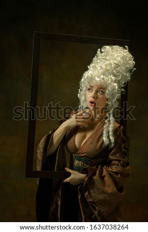 Astonished. Portrait of medieval young woman in vintage clothing with wooden frame on dark background. Female model as a duchess, royal person. Concept of comparison of eras, modern, fashion, beauty.