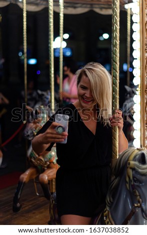 Portrait of a blonde woman take selfie with her smartphone on bright carousel at night in the city of Florence. Concept of travel, destination, summer, vacation.