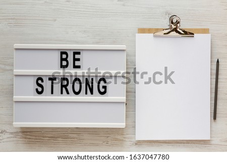 'Be strong' words on a lightbox, noticepad with blank sheet of paper on a white wooden background, top view. Overhead, from above, flat lay. 