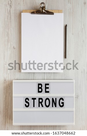 'Be strong' words on a modern board, noticepad with blank sheet of paper on a white wooden background, top view. Overhead, from above, flat lay. 