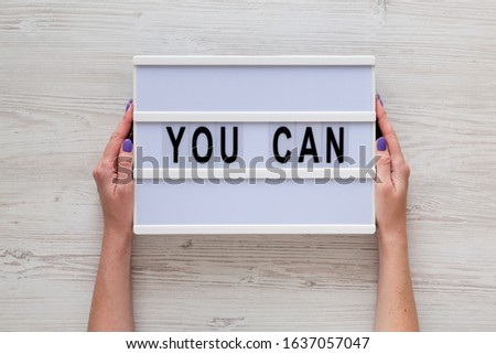 Female hands hold a lightbox with 'You can' words over white wooden surface, top view. Overhead, from above, flat lay. 